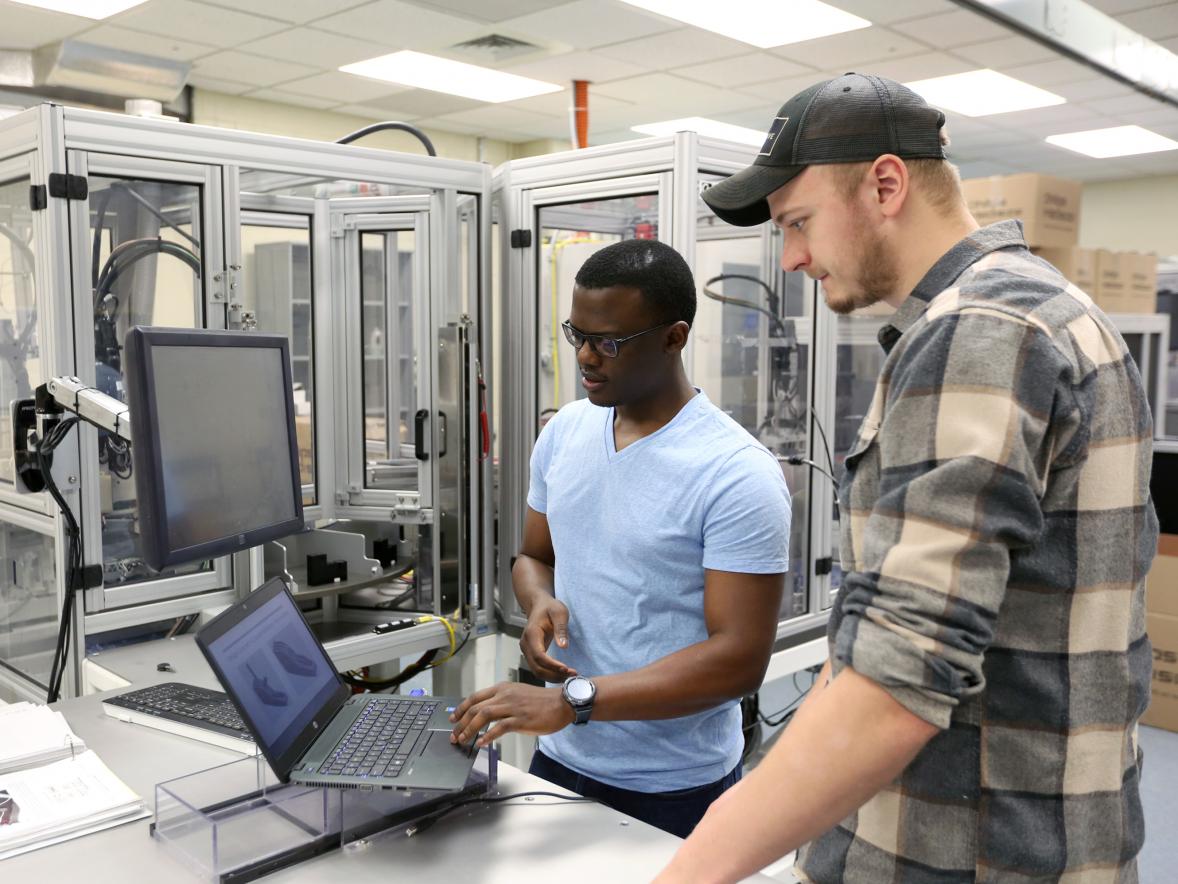 Students Kefa Okoth, left, and Ben Miner review plans to reconfigure a donated manufacturing cell, background, for a capstone engineering project. 